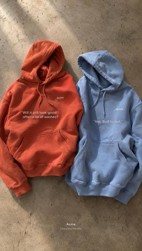 hoodies flat lay product photo: Two orange and blue hoodies lie side by side on a textured concrete surface, with small white copy on the garments and soft dir…