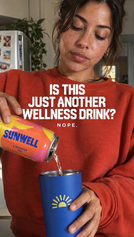 canned wellness drink splash / pour product photo: A woman in a warm red sweatshirt pours a brightly colored canned drink into a blue tumbler in a home kitchen…