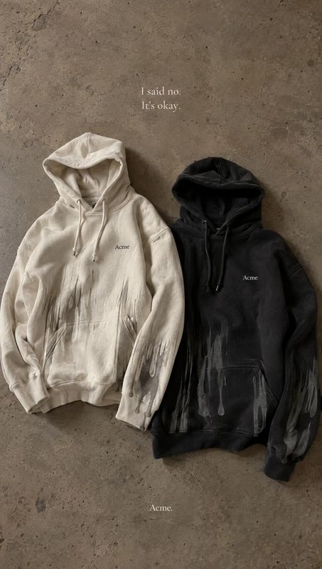 hoodies flat lay product photo: Two cream and black hoodies with painterly handprints lie on a textured concrete surface, styled with understated serif copy fo…