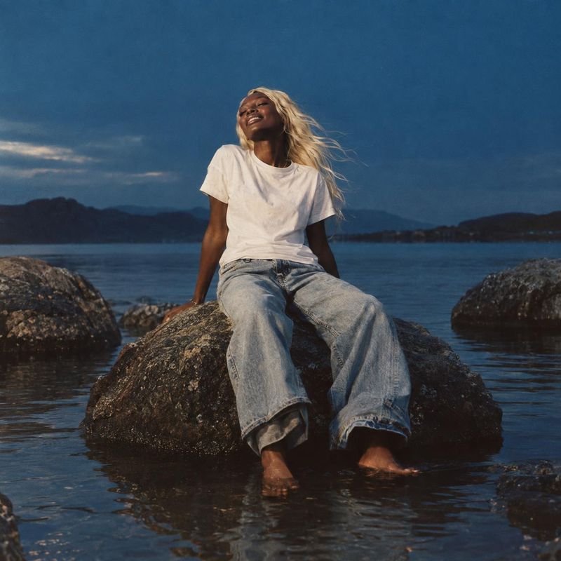 casual outfit action & motion product photo: A woman in a white T-shirt and loose light-wash jeans sits barefoot on a seaside rock, her hair lifted by the bree…