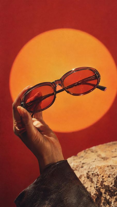 sunglasses product in hand product photo: A hand presents dark oval sunglasses against a vivid red backdrop with a glowing orange circle and warm directional l…