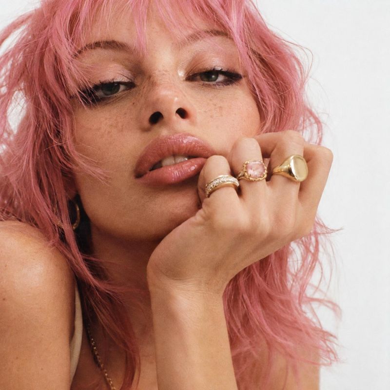 gold rings on-body detail product photo: A close-up studio portrait shows a woman with tousled pink hair, soft makeup and several gold rings posed against a pa…