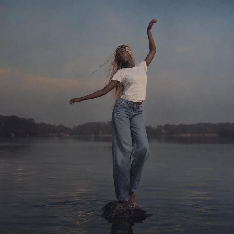 jeans and casual outfit action & motion product photo: A model in a white T-shirt and relaxed blue jeans balances on a rock in calm water, her arms raised in a…