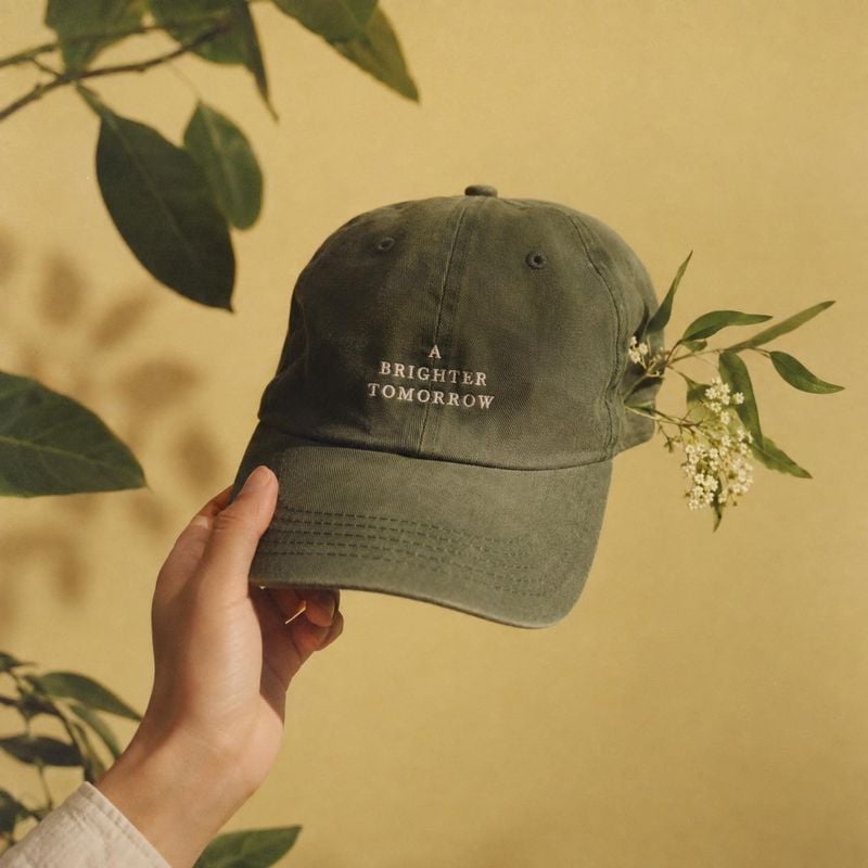 embroidered baseball cap product in hand product photo: A hand presents an olive cap embroidered with “A Brighter Tomorrow” against a warm beige backdrop, fram…