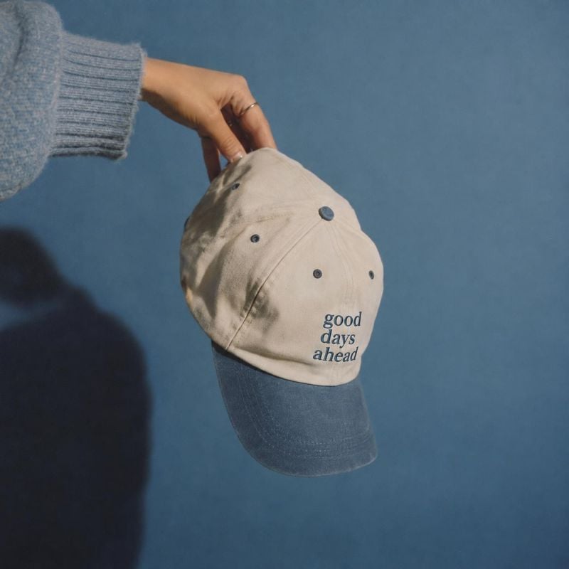 embroidered baseball cap product in hand product photo: A hand in a soft blue knit sleeve holds a beige-and-blue embroidered cap against a muted blue backdrop,…