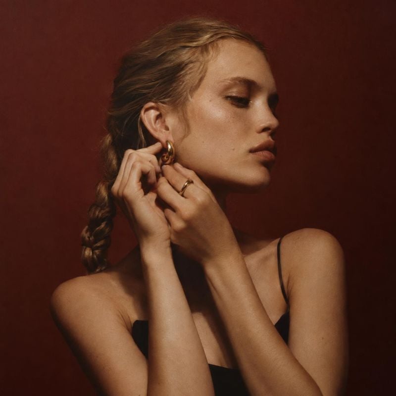 gold hoop earrings on-body detail product photo: A woman adjusts a gold hoop earring against a deep burgundy studio backdrop, with warm directional light and a…