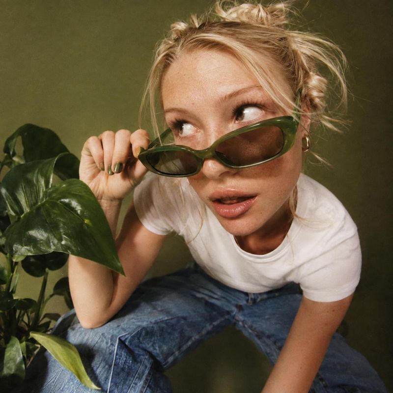 sunglasses on-body detail product photo: A blonde model in a white T-shirt and blue jeans lowers dark green sunglasses beside a leafy plant, photographed again…