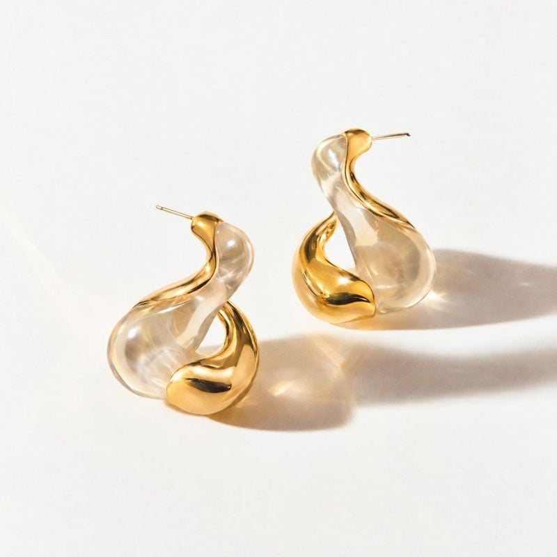sculptural gold-tone earrings jewelry product photo: A pair of sculptural gold-tone earrings with translucent curved forms rests on a bright white surface, lit…