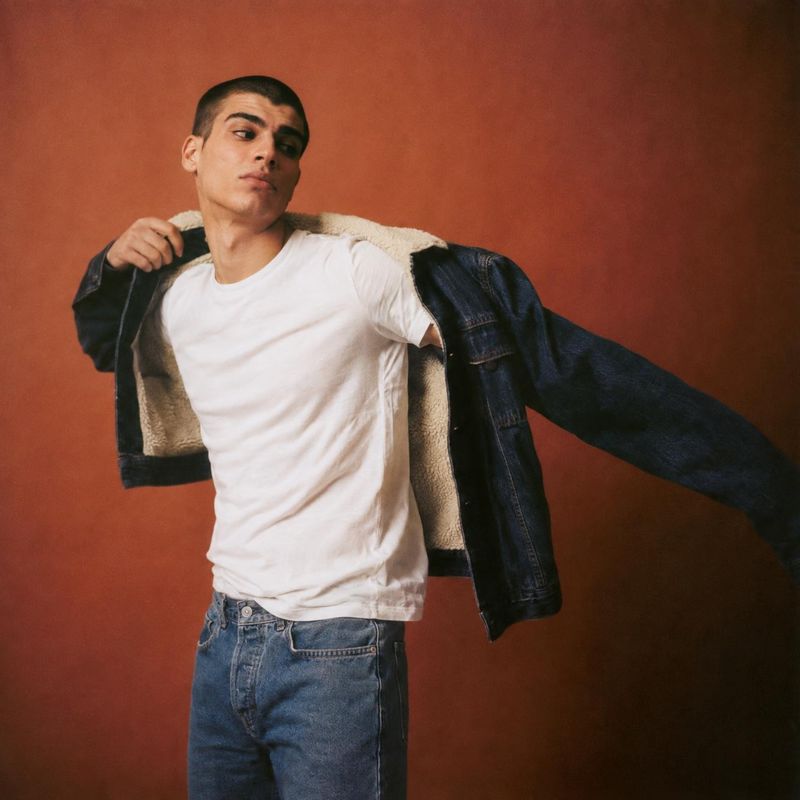 denim jacket action & motion product photo: A male model opens a shearling-lined denim jacket over a white T-shirt and jeans against a warm terracotta studio b…