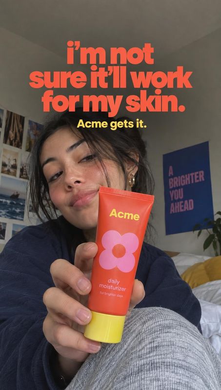 daily moisturizer product in hand product photo: A woman presents a bright coral moisturizer tube toward the camera in a casual bedroom selfie, with bold coral…
