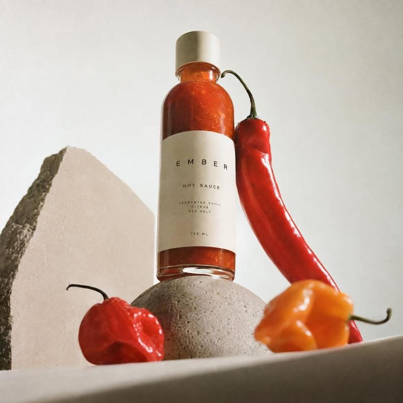 hot sauce ingredients product photo: A bottle of amber-red hot sauce stands among fresh red and orange chilies on sculptural stone forms, lit softly against a…