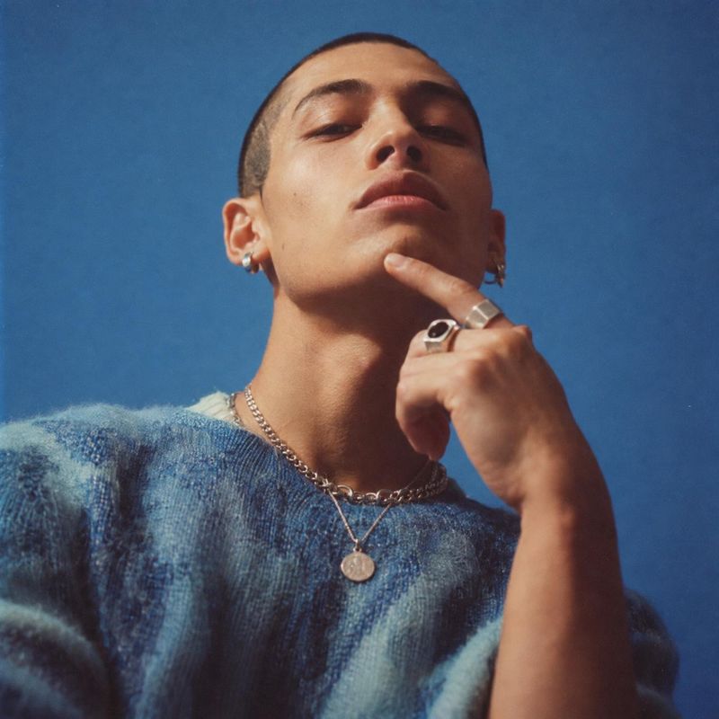 silver jewelry and knitwear on-body detail product photo: A low-angle fashion portrait shows a model in a blue knit sweater, layered silver necklaces, earrings…
