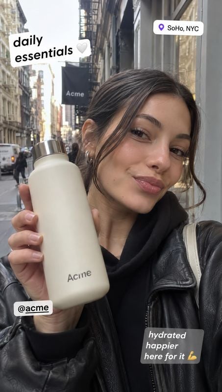 reusable water bottle product in hand product photo: A woman in a black leather jacket holds a cream-colored bottle on a SoHo street, with casual social-media…