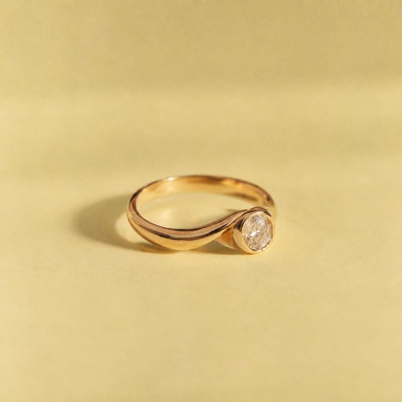 gold diamond ring jewelry product photo: A gold ring with a single round clear stone rests on a warm yellow seamless surface, lit softly to create a refined, m…