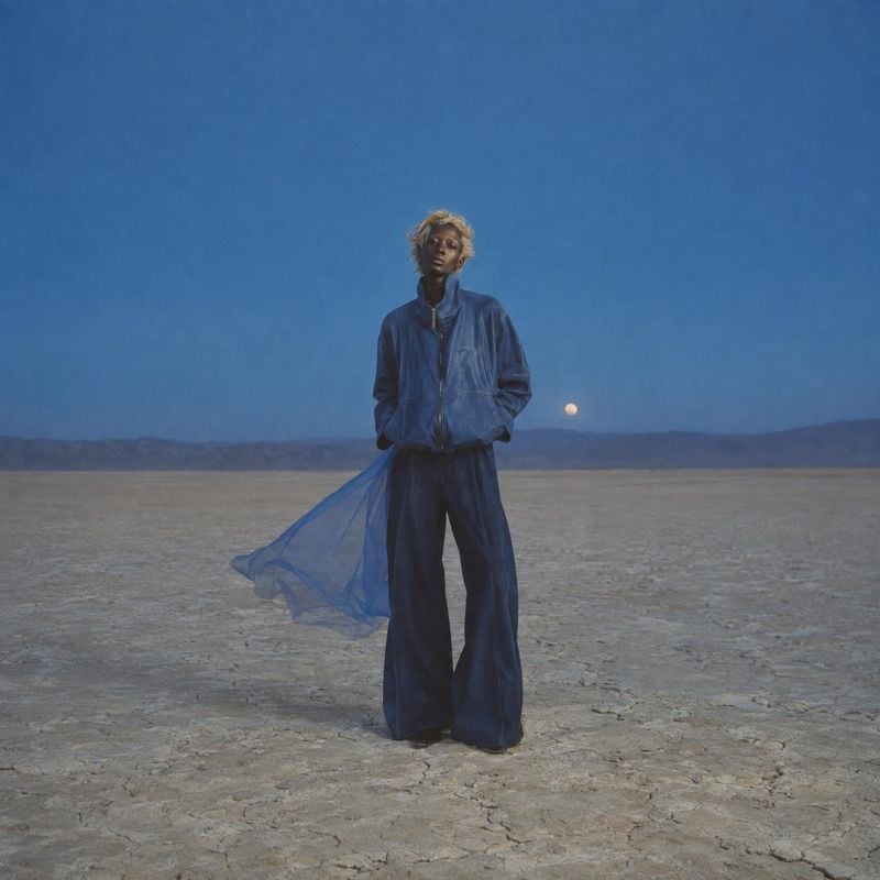 blue jacket and wide-leg trousers action & motion product photo: A model in an oversized blue jacket and wide-leg trousers stands on a barren salt flat, with s…
