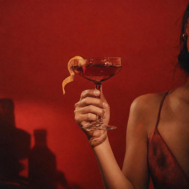 cocktail product in hand product photo: A woman in a red slip dress holds a coupe of red cocktail garnished with citrus against a deep red backdrop, creating a…