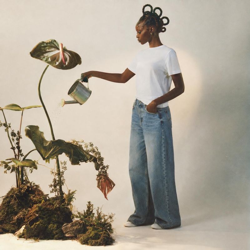 jeans and T-shirt equipment in use product photo: A model in a white T-shirt and relaxed blue jeans waters a sculptural arrangement of leafy plants against a w…