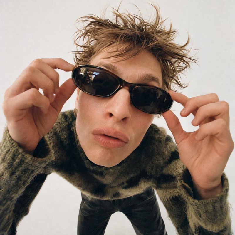 sunglasses on-body detail product photo: A tousle-haired male model leans toward the camera while adjusting dark sunglasses, styled with a chunky patterned sca…