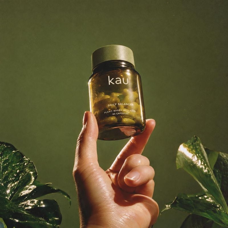 wellness supplement capsules product in hand product photo: A hand presents an amber bottle of wellness capsules against a deep green backdrop with glossy leav…
