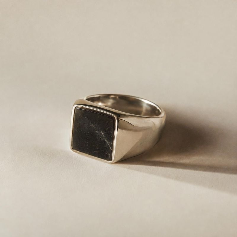 silver signet ring jewelry product photo: A polished silver signet ring with a dark square stone rests on a warm neutral surface, lit softly from the side for…