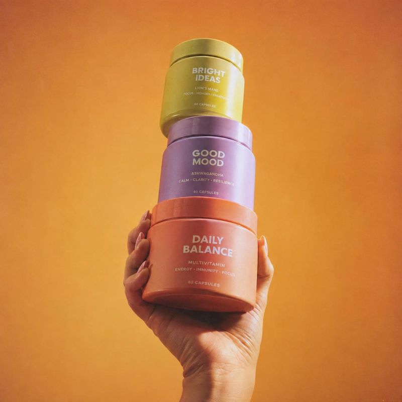 stack of supplement bottles product in hand product photo: A hand presents a colorful stack of supplement bottles against a warm orange studio backdrop, creati…