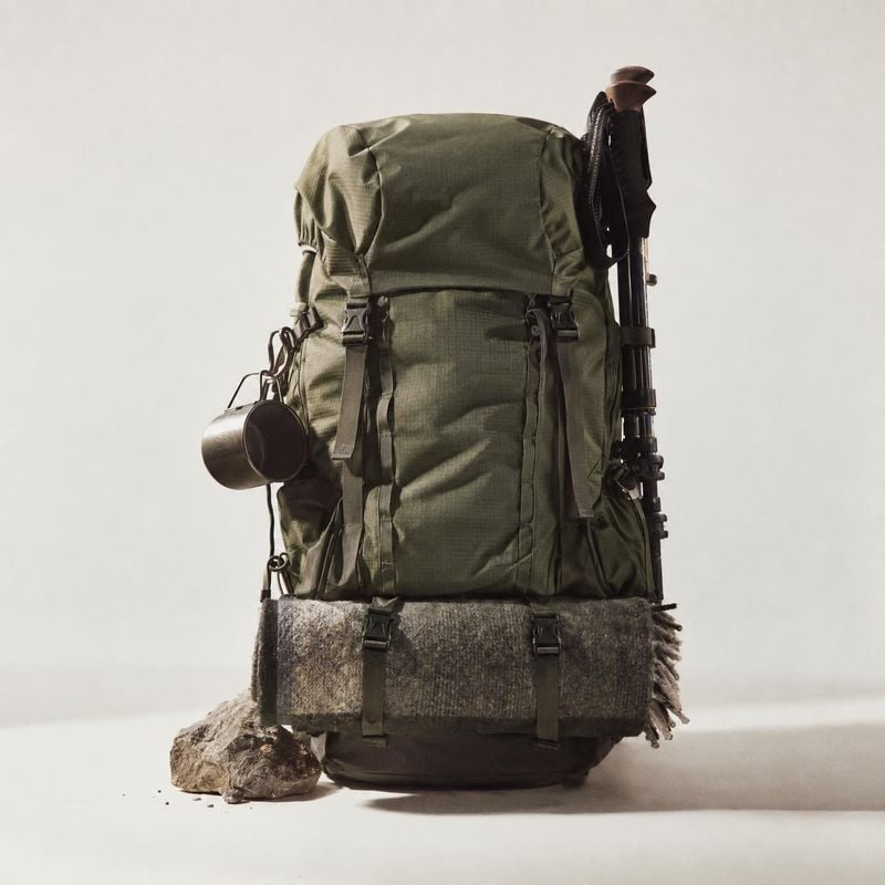 hiking backpack sports product photo: An olive hiking backpack fitted with a blanket, cup and trekking gear stands against a softly lit neutral studio backdrop…