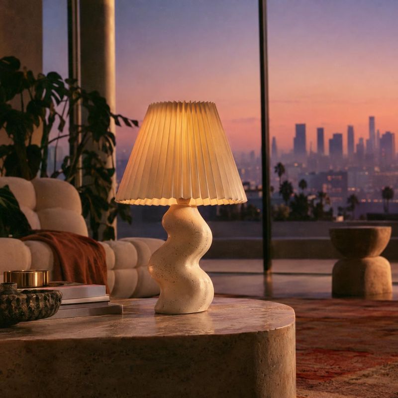 table lamp lifestyle scene product photo: A sculptural cream table lamp glows on a stone coffee table in a softly furnished living room, with a dusky city skyl…