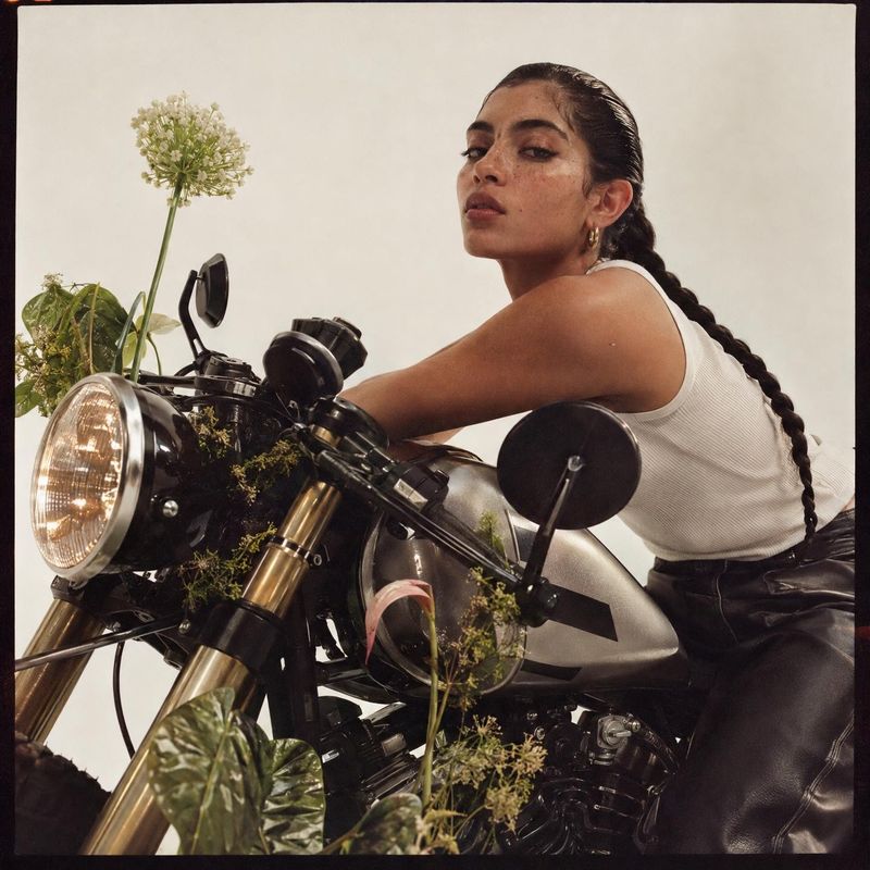 motorcycle vehicle exterior product photo: A woman in a white ribbed top and black leather pants poses astride a dark motorcycle among leafy plants against a p…