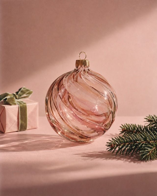 glass Christmas ornament christmas product photo: A pink twisted-glass Christmas ornament hangs above a matching gift box and evergreen sprig on a blush-pink s…