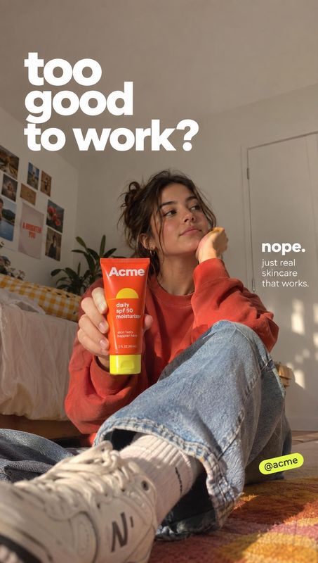 sunscreen moisturizer lifestyle scene product photo: A young woman in a red sweatshirt sits casually in a sunlit bedroom, holding a bright sunscreen tube towar…