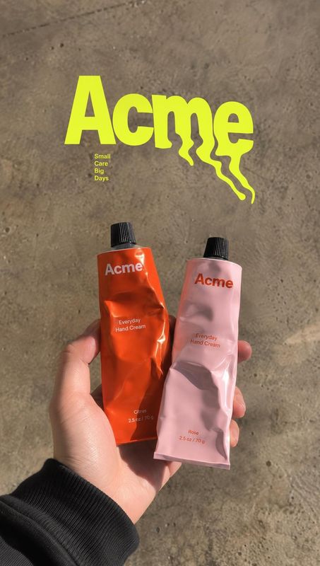 hand cream product in hand product photo: A hand holds orange and pink hand-cream tubes against rough concrete, with oversized neon-yellow lettering adding a b…