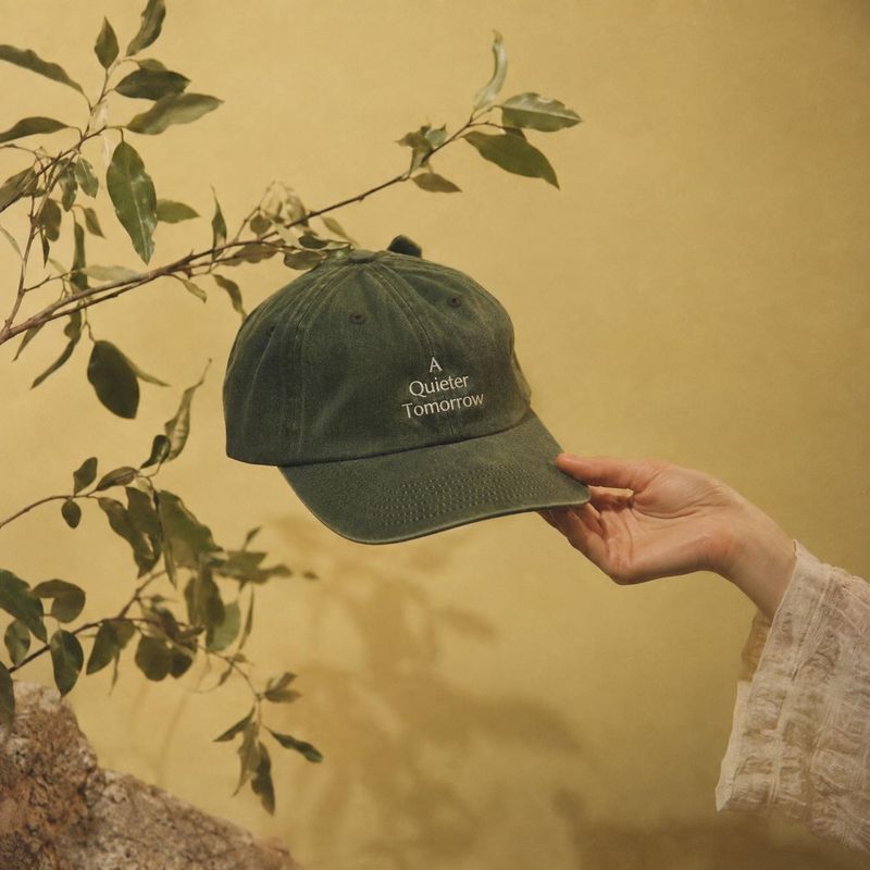 baseball cap product in hand product photo: A hand presents a dark green embroidered cap against a warm ochre backdrop, framed by leafy branches in soft, earth…