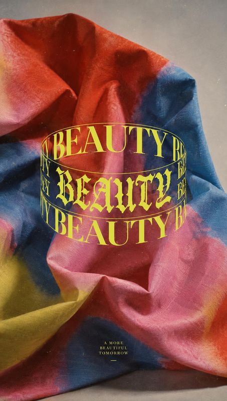 close-up detail product photo: A vivid red, blue, and yellow fabric is artfully draped beneath oversized gold “BEAUTY” typography, with a small hopeful tagline…
