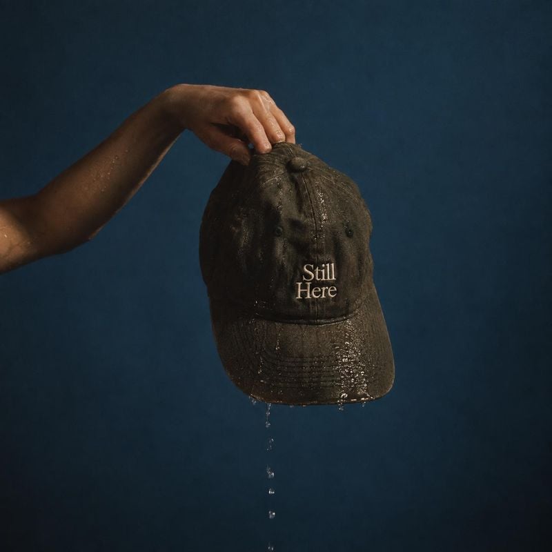 baseball cap product in hand product photo: A hand holds a dripping, weathered dark cap against a deep blue studio backdrop, with dramatic directional light an…