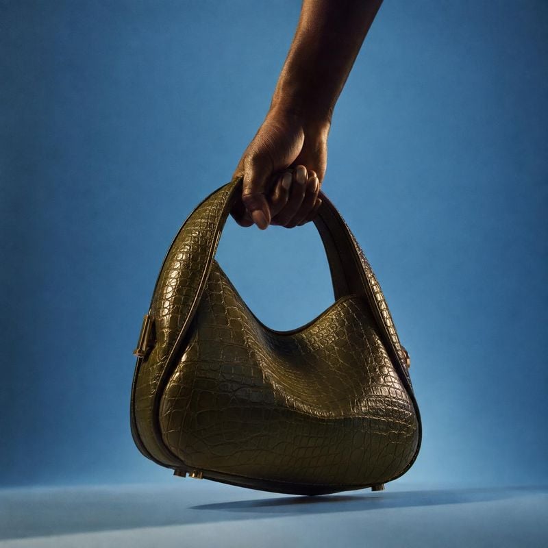 handbag product in hand product photo: A dark olive crocodile-textured handbag is held by its handle against a blue studio backdrop, with dramatic directional…