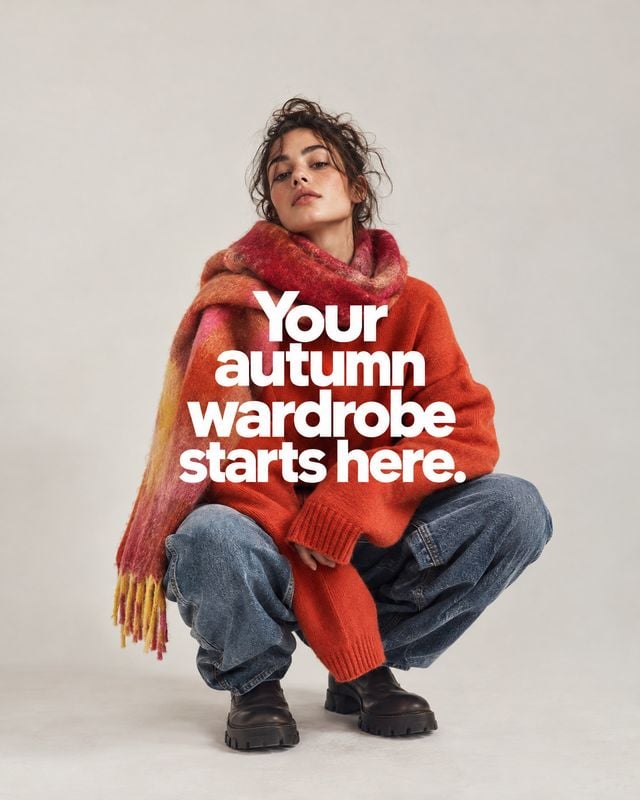 autumn outfit autumn product photo: A seated model wears a vivid red sweater, multicolored scarf, jeans, and chunky boots against a pale studio backdrop, with…