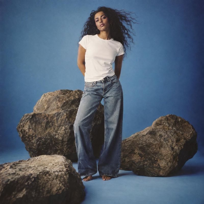 jeans and T-shirt action & motion product photo: A model poses in a white T-shirt and relaxed blue jeans among rugged rocks against a blue studio backdrop, wit…
