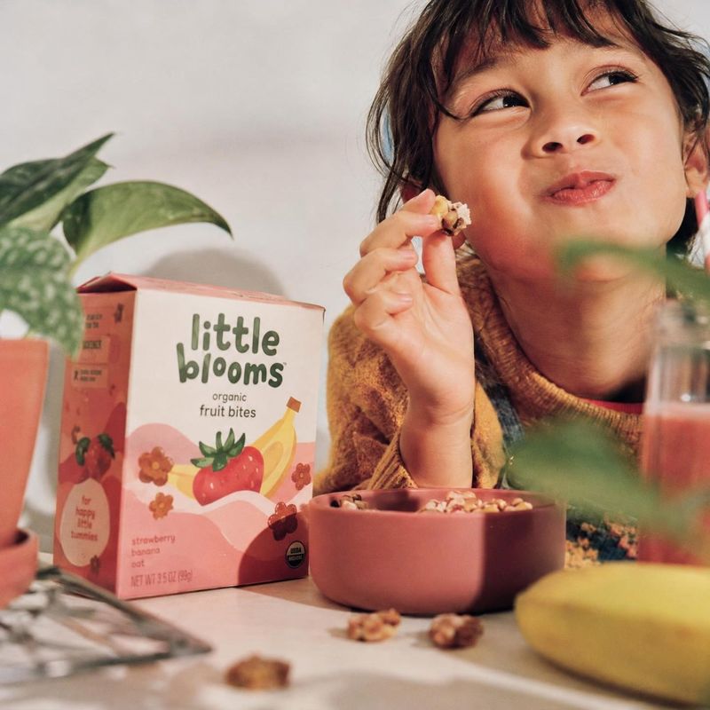 organic fruit bites lifestyle scene product photo: A child enjoys a bite-sized snack beside its illustrated fruit-bite box and a pink bowl, in a warm, softly l…