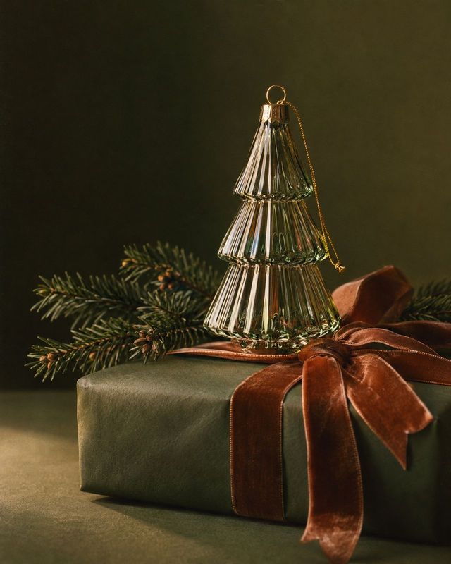 glass Christmas tree ornament christmas product photo: A tiered glass tree ornament rests atop a dark green gift wrapped with a rust velvet bow and evergreen s…