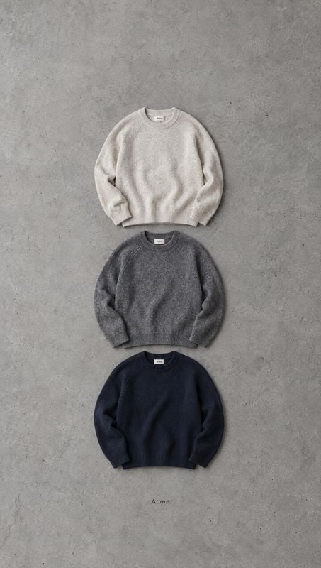 sweaters flat lay product photo: Three knit sweaters in cream, charcoal, and navy are arranged vertically on a textured gray concrete surface, with soft even l…