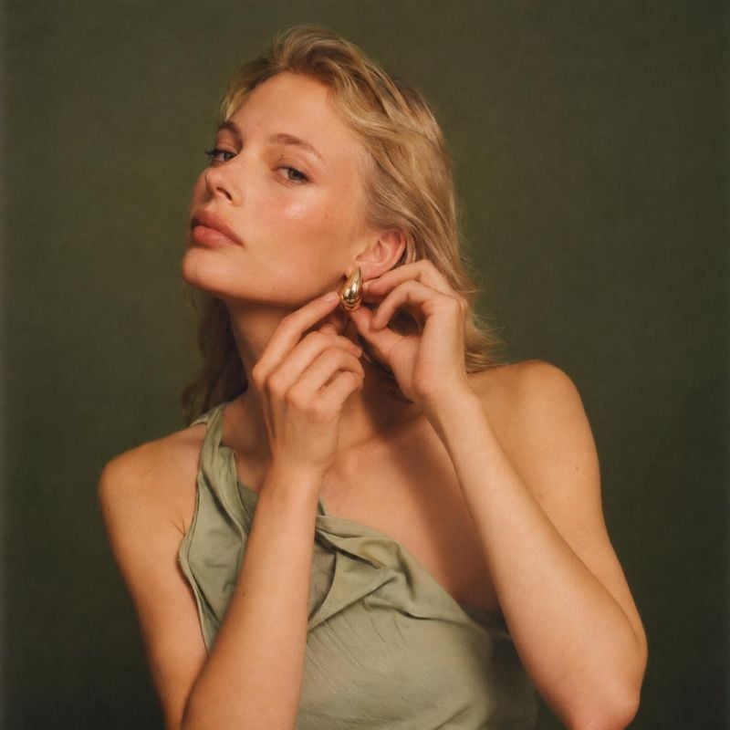 gold statement earring on-body detail product photo: A blonde model adjusts a sculptural gold earring against a muted olive studio backdrop, with warm soft lig…