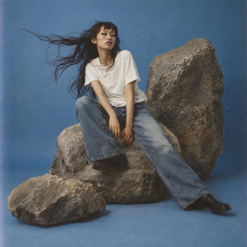 wide-leg jeans action & motion product photo: A model in a white T-shirt, wide-leg jeans and dark shoes sits among rugged rocks against a blue studio backdrop,…