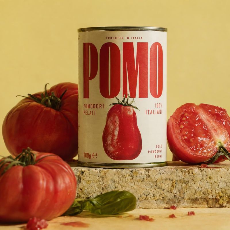 peeled tomato can ingredients product photo: A can of peeled tomatoes stands among ripe whole and halved tomatoes on a stone surface, styled against a warm yel…