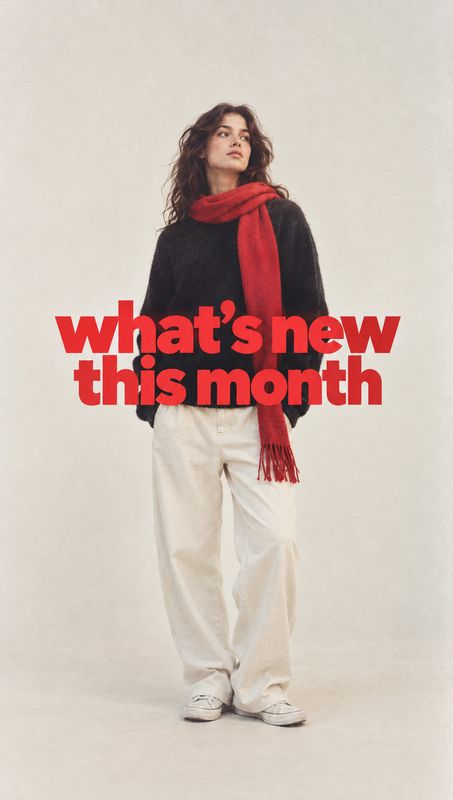 fashion outfit and scarf black friday product photo: A model in a black sweater, red scarf, and loose white trousers poses against a softly lit neutral studio…