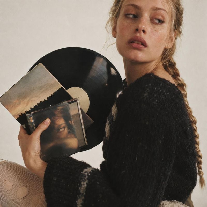 vinyl record and CD book product photo: A freckled model in a chunky black sweater holds a vinyl record and CD against a softly lit neutral studio backdrop, cr…