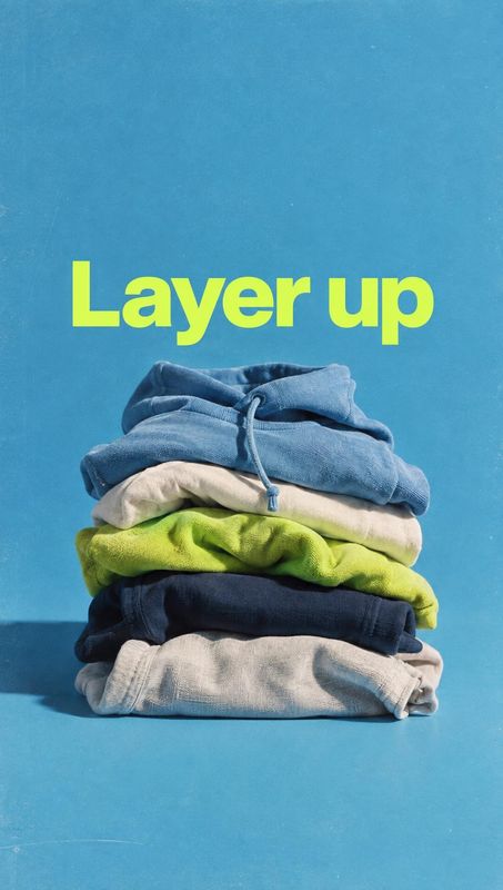 stack of folded hoodies fashion product photo: A stack of colorful folded hoodies sits against a bright blue studio backdrop beneath a bold lime-green “Layer u…