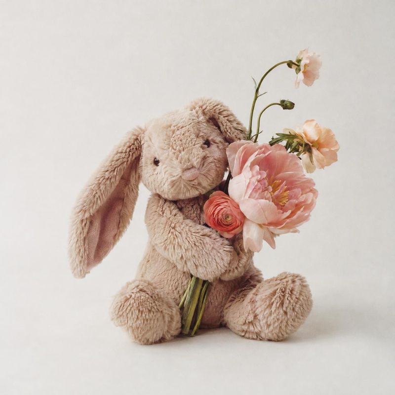 plush bunny toy toys product photo: A soft beige plush bunny cradles pink flowers against a pale seamless background, styled with gentle natural light for a sw…