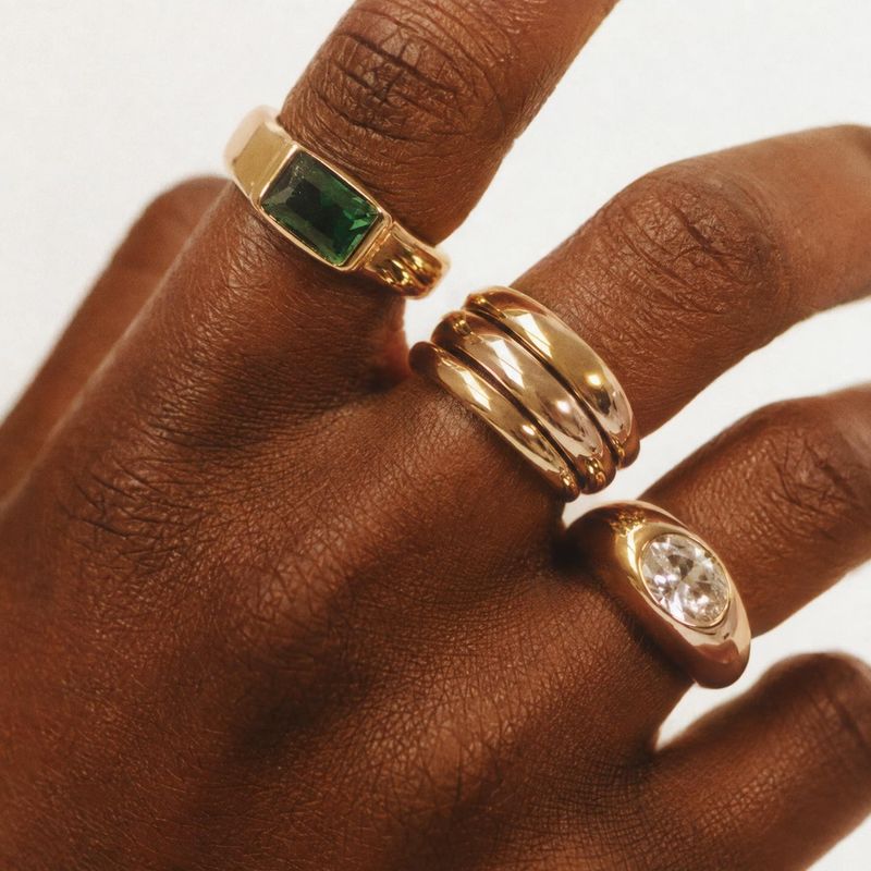 gold rings on-body detail product photo: A close-up of several polished gold rings, including green and clear stones, worn on a hand against a bright neutral b…