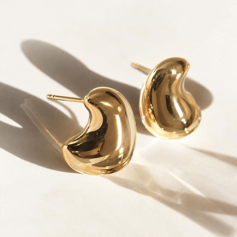 gold-tone stud earrings flat lay product photo: A pair of glossy, sculptural gold-tone stud earrings rests on a warm ivory surface, lit by direct sunlight that…