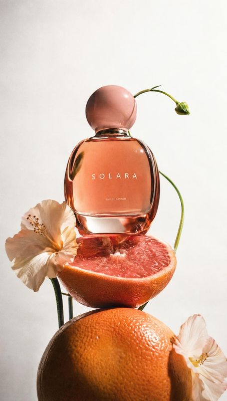 perfume ingredients product photo: A peach-toned perfume bottle is styled with grapefruit and delicate flowers against a softly lit, pale background, creating…